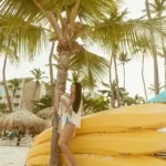 A woman relaxes by a palm tree on a sunny beach with yellow kayaks nearby.