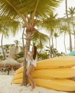 A woman relaxes by a palm tree on a sunny beach with yellow kayaks nearby.