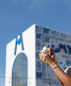 A person holds a skincare product against a colorful building with a blue sky backdrop.