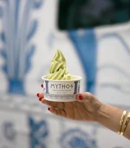 A hand holds a cup of Mythos Greek frozen yogurt, showcasing a creamy swirl of delicious treat.